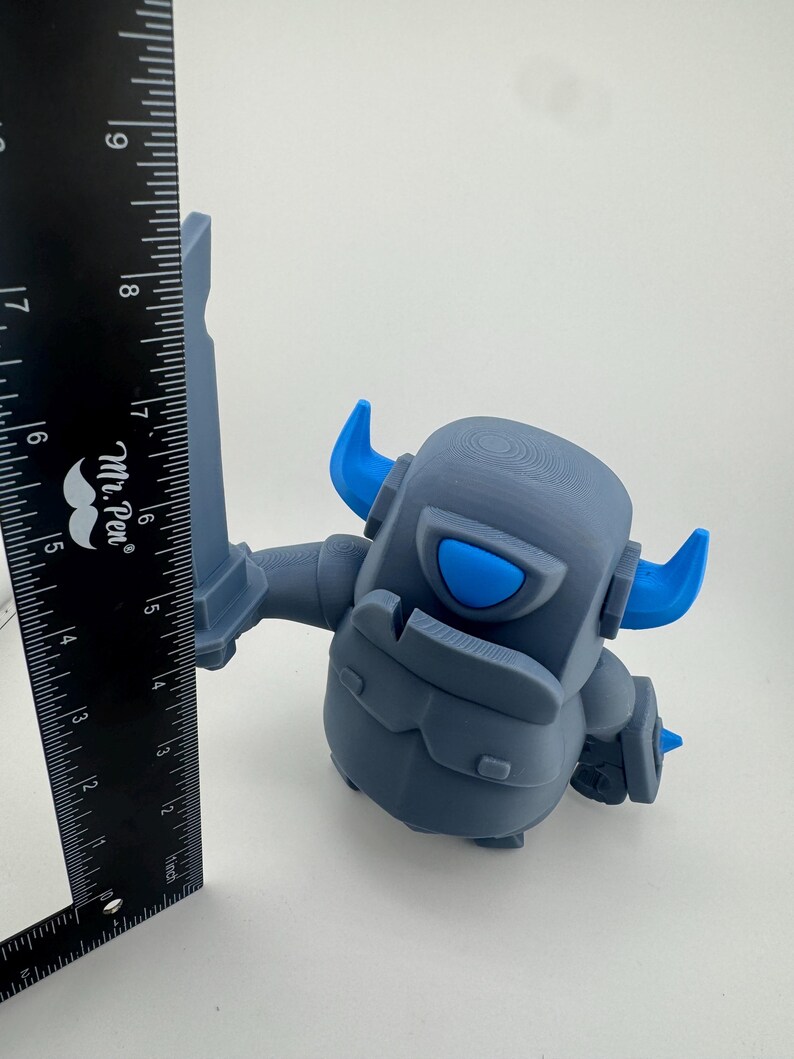 May include: A gray and blue plastic figurine of a knight character holding a sword, positioned next to a black Mr. Pen ruler. The ruler shows the figurine stands approximately 5.5 inches tall from the base to the top of the sword blade.