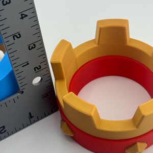 Puede incluir: Objeto impreso en 3D con forma de corona, con interior rojo y exterior dorado. La corona tiene una base circular y mide aproximadamente 7,6 cm de di&aacute;metro.
