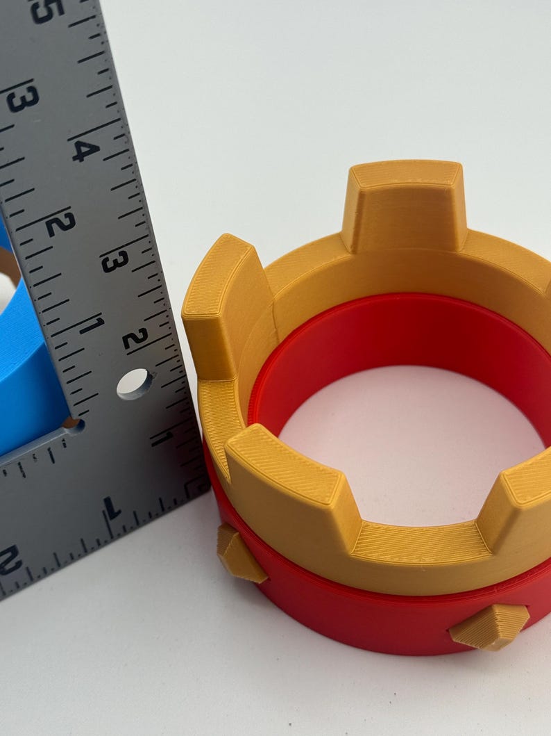 Puede incluir: Objeto impreso en 3D con forma de corona, con interior rojo y exterior dorado. La corona tiene una base circular y mide aproximadamente 7,6 cm de di&aacute;metro.