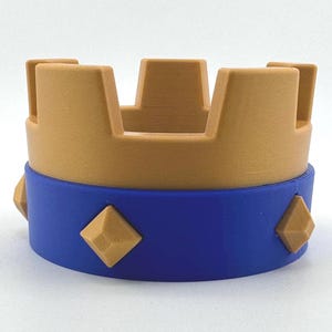 Puede incluir: Una corona impresa en 3D en dos tonos. La parte superior es de color beige, con cortes rectangulares. La banda inferior es azul, adornada con cuatro acentos en forma de diamante de color beige. Un objeto decorativo.