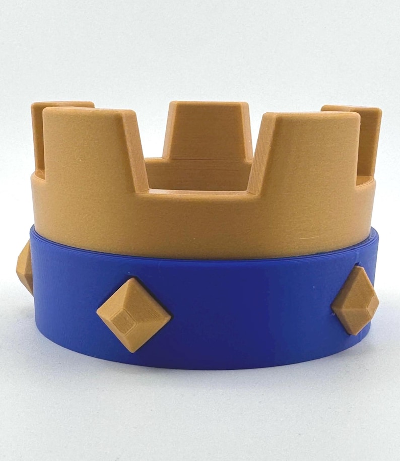 Puede incluir: Una corona impresa en 3D en dos tonos. La parte superior es de color beige, con cortes rectangulares. La banda inferior es azul, adornada con cuatro acentos en forma de diamante de color beige. Un objeto decorativo.