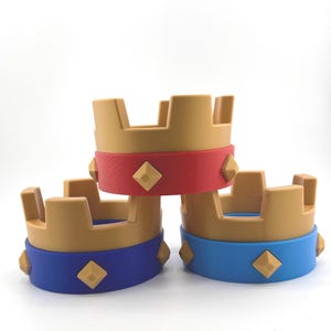 Puede incluir: Tres coronas de juguete apiladas. Las coronas tienen una parte superior de color canela y una banda roja, azul y azul. Cada banda tiene cuatro acentos en forma de diamante. El fondo es blanco.