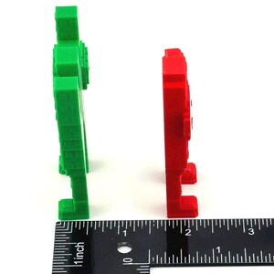 May include: Two pixelated figures, one green and one red, stand side-by-side against a white background. A ruler shows the figures are approximately 2 inches tall. The figures have a blocky, retro video game aesthetic.