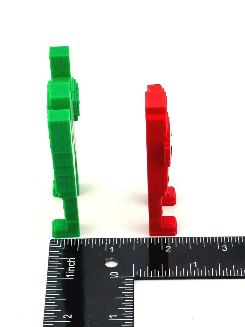 May include: Two pixelated figures, one green and one red, stand side-by-side against a white background. A ruler shows the figures are approximately 2 inches tall. The figures have a blocky, retro video game aesthetic.