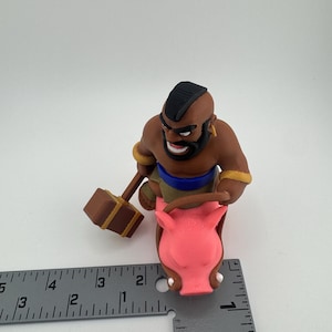 Puede incluir: Figura impresa en 3D de un personaje montado en un cerdo rosa. El personaje tiene una cresta negra, una barba marr&oacute;n y sostiene un martillo marr&oacute;n. El cerdo tiene un hocico y orejas detallados. La figura mide aproximadamente 5 cm de altura.