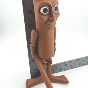 May include: A brown, articulated figurine with large eyes and a smiling expression. The figure is approximately 7 inches tall, holding a brown stick. The figure is standing on a white surface.