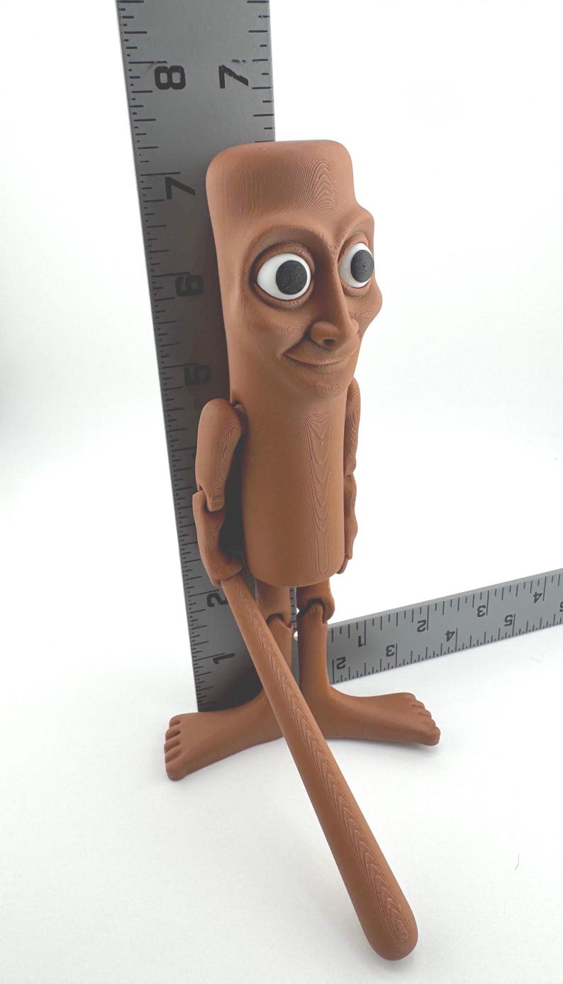 May include: A brown, articulated figurine with large eyes and a smiling expression. The figure is approximately 7 inches tall, holding a brown stick. The figure is standing on a white surface.
