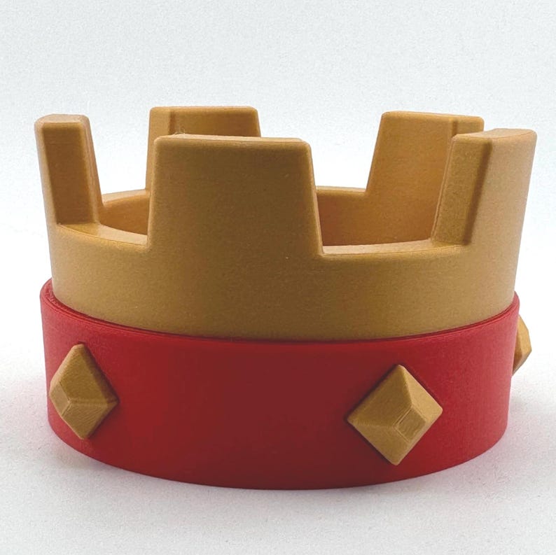 Puede incluir: Una corona dorada con una banda roja. La corona tiene una serie de formas rectangulares verticales. La banda roja presenta tres detalles dorados en forma de diamante. La corona es un objeto decorativo.