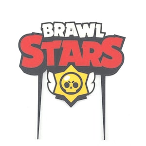 May include: A cake topper featuring the words "BRAWL STARS" in white and red, with a black outline. Below the text is a yellow star with a skull icon. The topper has two black prongs for insertion into a cake.
