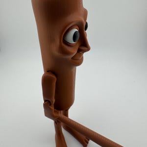 May include: A brown, articulated figurine with a rectangular head, large eyes, and a smiling expression. The figure has jointed arms and legs, and is holding a long, brown, cylindrical object. The figurine is standing on a white surface.