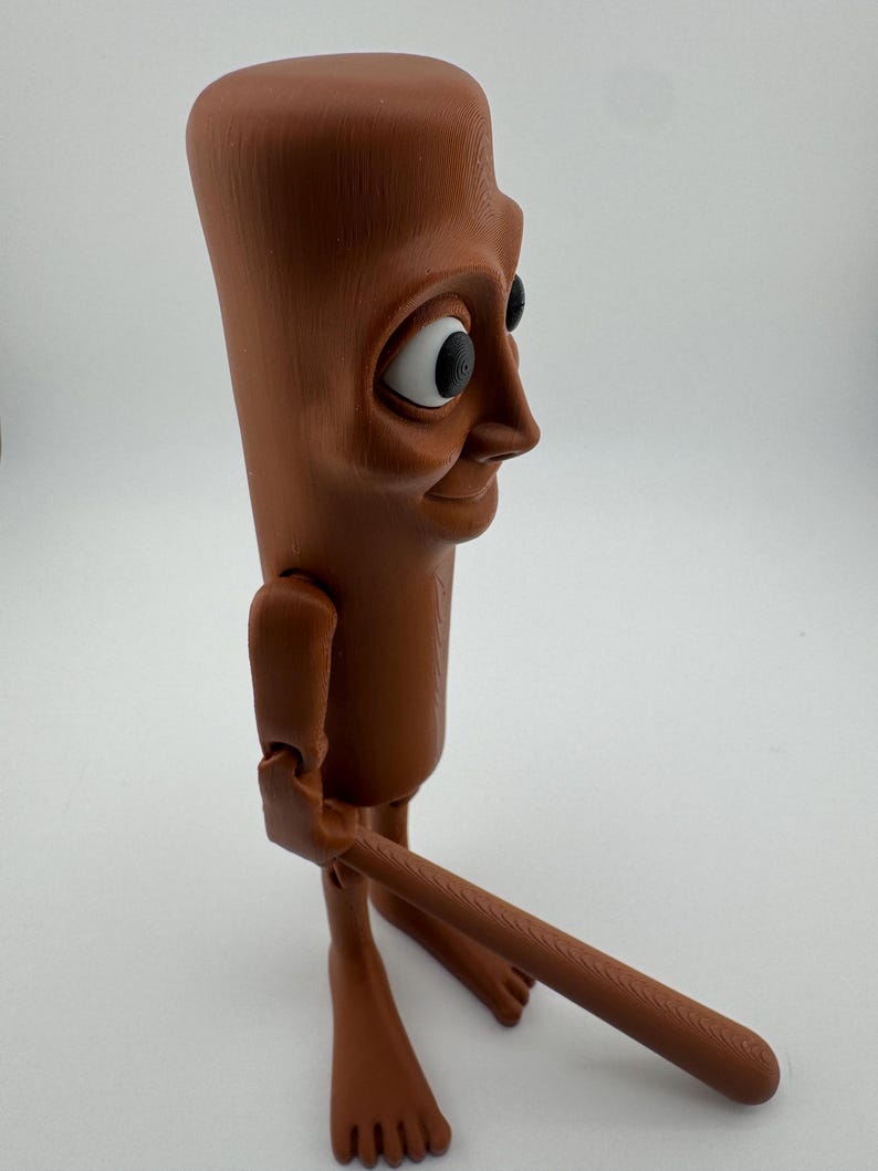 May include: A brown, articulated figurine with a rectangular head, large eyes, and a smiling expression. The figure has jointed arms and legs, and is holding a long, brown, cylindrical object. The figurine is standing on a white surface.