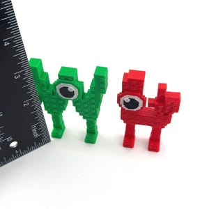 May include: Two pixelated figures, one green and one red, each with a large eye. The green figure is taller and resembles the letter "W". The red figure is shorter and has a more rectangular shape. A ruler is placed next to the figures.