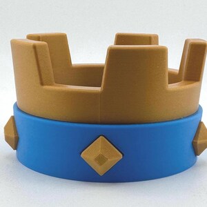 Puede incluir: Una corona de juguete con la parte superior dorada y una base azul. La corona tiene cuatro secciones rectangulares verticales y est&aacute; decorada con formas de diamante doradas. La base tiene una banda azul con detalles en forma de diamante dorados.