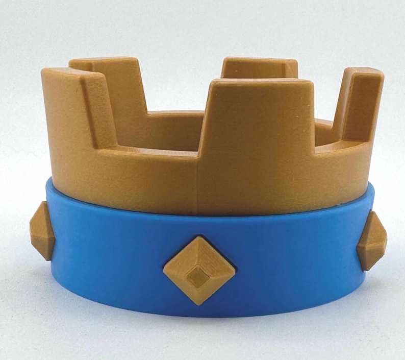 Puede incluir: Una corona de juguete con la parte superior dorada y una base azul. La corona tiene cuatro secciones rectangulares verticales y est&aacute; decorada con formas de diamante doradas. La base tiene una banda azul con detalles en forma de diamante dorados.