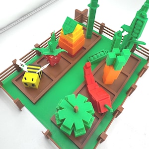 May include: A colorful 3D-printed garden scene featuring various vegetables and a bee. The garden includes a strawberry, a carrot, a pepper, and a green leafy plant, all arranged within a brown-fenced plot on a green base.