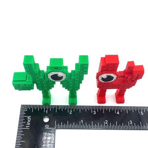 May include: Two pixelated creature figurines. One is green, the other is red. Both have large, white-eyed faces. A ruler is placed below the figurines to show their size.