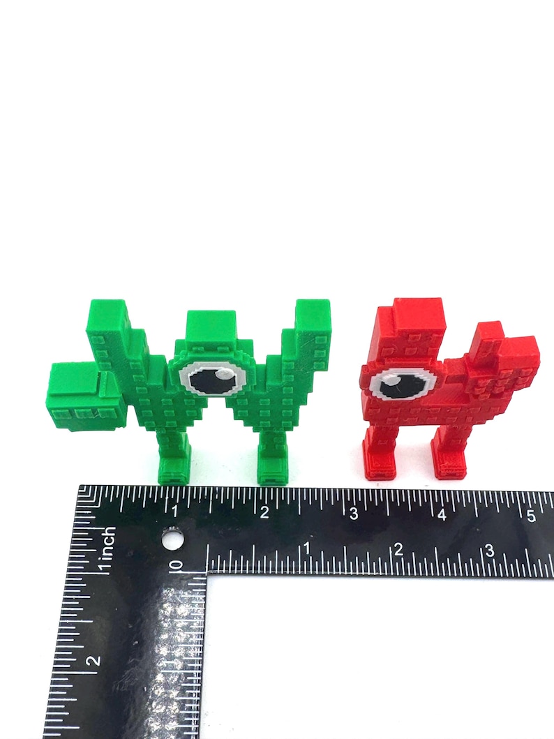 May include: Two pixelated creature figurines. One is green, the other is red. Both have large, white-eyed faces. A ruler is placed below the figurines to show their size.