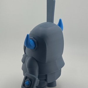 May include: A gray 3D printed figurine of a knight character with a large sword extending from the back. The figure features two bright blue horns on the helmet and a small blue accent on the shield held in the hand, with a smooth matte finish.