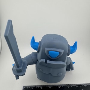 May include: A gray 3D-printed figurine of a warrior character with bright blue horns and a blue eye visor, holding a gray sword. A black ruler is positioned at the base, showing the figure stands approximately 4.5 inches in total height.