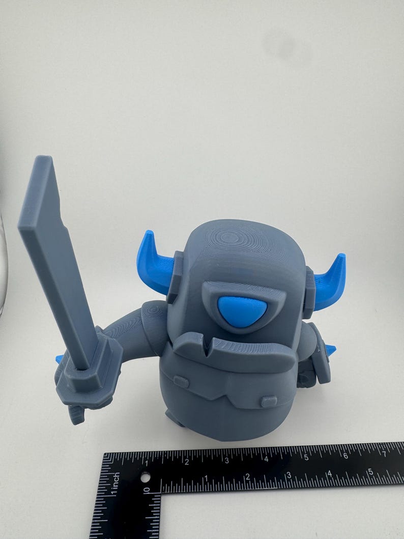 May include: A gray 3D-printed figurine of a warrior character with bright blue horns and a blue eye visor, holding a gray sword. A black ruler is positioned at the base, showing the figure stands approximately 4.5 inches in total height.