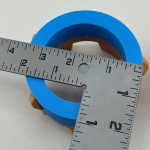 Puede incluir: Un objeto en forma de anillo impreso en 3D, azul y marr&oacute;n. El objeto se mide con una regla. El anillo azul tiene un di&aacute;metro exterior de aproximadamente 7,6 cm. La parte marr&oacute;n es visible en los lados del anillo.
