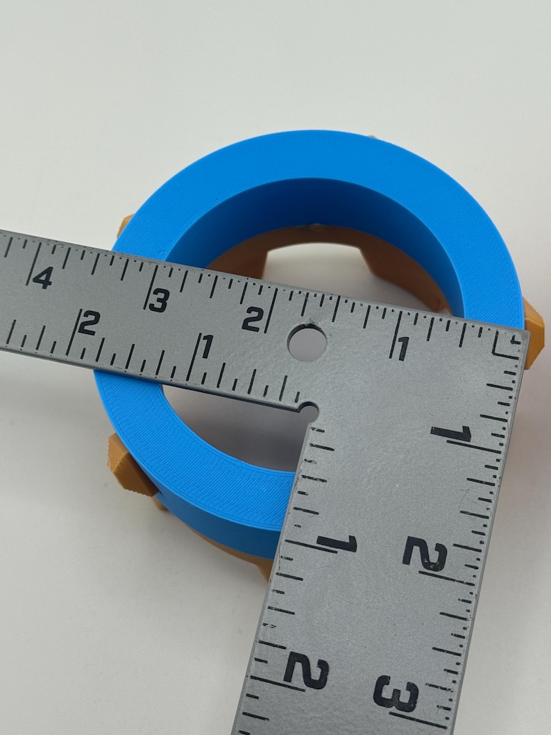 Puede incluir: Un objeto en forma de anillo impreso en 3D, azul y marr&oacute;n. El objeto se mide con una regla. El anillo azul tiene un di&aacute;metro exterior de aproximadamente 7,6 cm. La parte marr&oacute;n es visible en los lados del anillo.