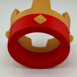 Puede incluir: Una corona impresa en 3D en oro y rojo. La corona presenta una banda roja alrededor de la base, con detalles dorados y adornos en forma de diamante. La corona est&aacute; dise&ntilde;ada con una base circular y una secci&oacute;n central vertical.