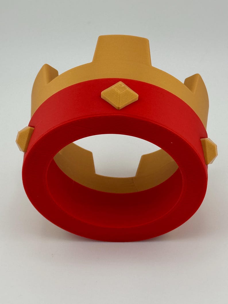Puede incluir: Una corona impresa en 3D en oro y rojo. La corona presenta una banda roja alrededor de la base, con detalles dorados y adornos en forma de diamante. La corona est&aacute; dise&ntilde;ada con una base circular y una secci&oacute;n central vertical.
