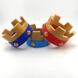 Puede incluir: Tres coronas de juguete con la parte superior dorada y una banda de color en la base. Una corona tiene una banda azul con detalles cuadrados azules, otra tiene una banda roja con detalles cuadrados rojos y la tercera tiene una banda azul.