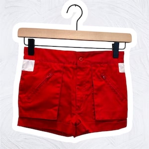 May include: Bright red shorts with white accents and two zippered pockets. The shorts are hanging on a wooden hanger with a black hook. The shorts have a button closure and a folded hem.