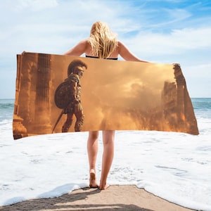 Puede incluir: Una toalla de playa rectangular con un guerrero con armadura griega antigua, sobre un fondo de ruinas y un cielo ardiente. La toalla la sostiene una persona de pie en una playa de arena con el océano al fondo.