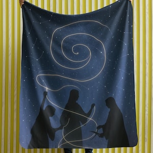 May include: A dark blue blanket featuring a starry night sky design with a swirling golden light pattern. Silhouettes of three figures are depicted below the spiral, holding the golden light. The blanket is held up against a yellow and white striped background.