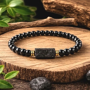 May include: Black beaded bracelet featuring a rectangular black stone and gold-coloured accents. The bracelet is made of small, round, glossy black beads. The bracelet is displayed on a wooden surface.