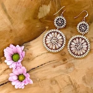 May include: A pair of dangle earrings with two circular beaded designs. The larger circle features a white and brown beaded pattern, while the smaller circle is brown. The earrings are hanging on a wooden surface.