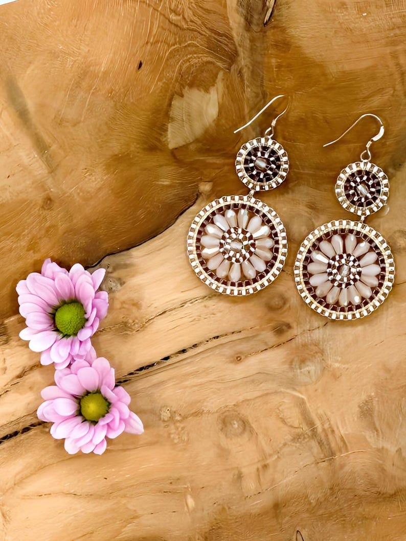 May include: A pair of dangle earrings with two circular beaded designs. The larger circle features a white and brown beaded pattern, while the smaller circle is brown. The earrings are hanging on a wooden surface.