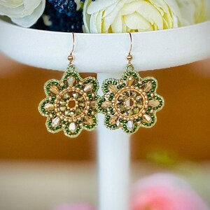 May include: A pair of gold and green beaded earrings with a floral design. The earrings are hanging from a white stand.
