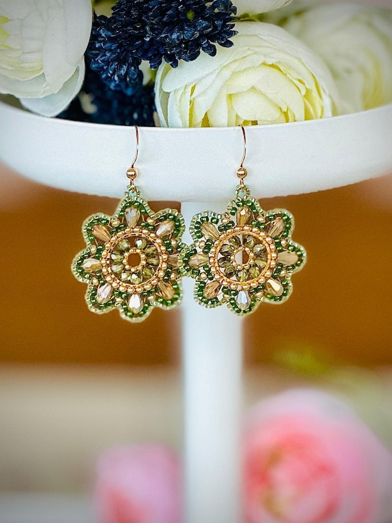 May include: A pair of gold and green beaded earrings with a floral design. The earrings are hanging from a white stand.