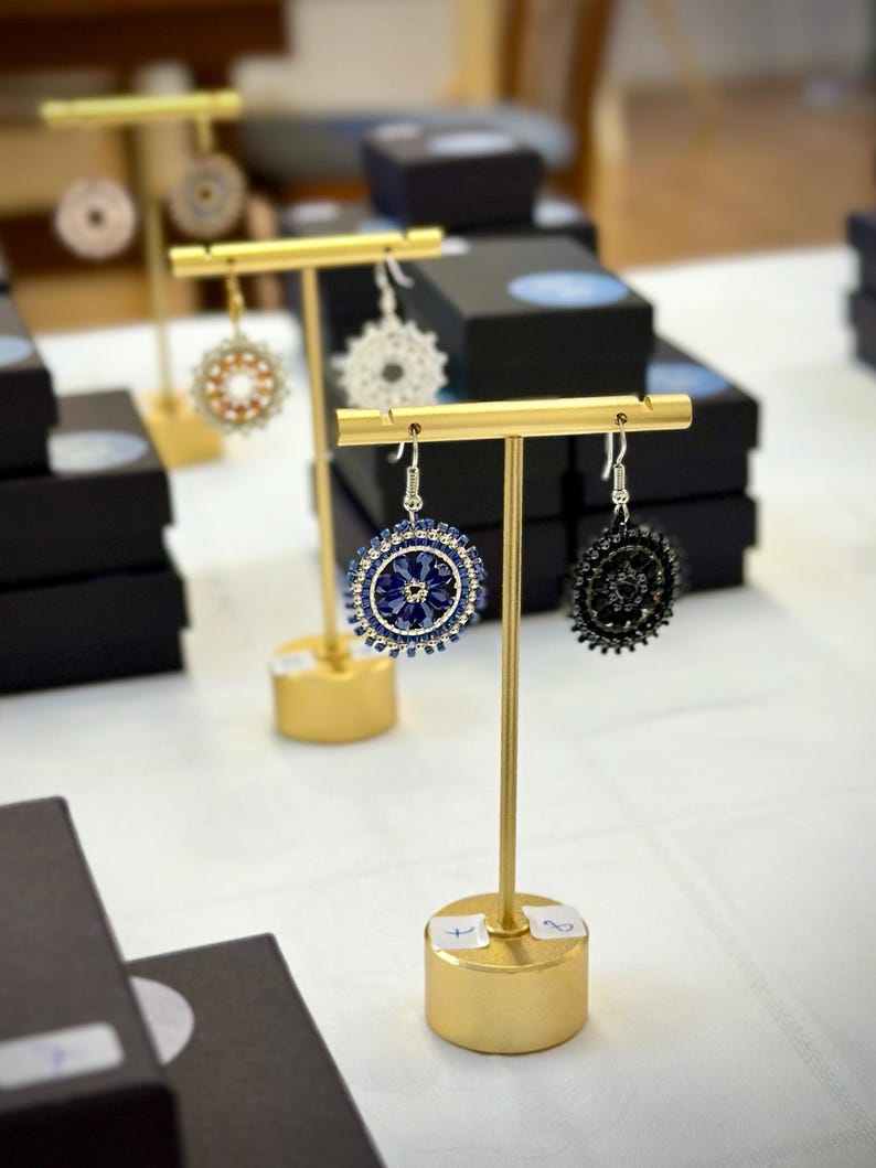 May include: Gold earring stands displaying a variety of earrings. The earrings are made of beads and feature different colours and patterns. The stands are on a white surface.