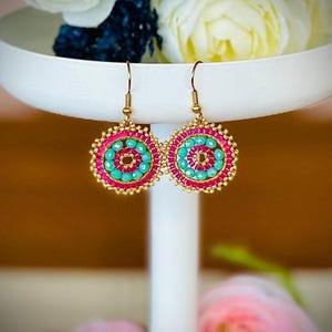 May include: A pair of gold hoop earrings with pink and turquoise beads. The earrings are on a white stand.