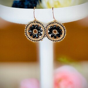 May include: A pair of gold and black beaded earrings with a flower design. The earrings are round and have a black centre with gold beads surrounding it.