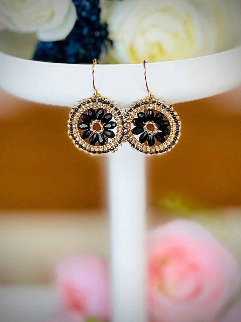 May include: A pair of gold and black beaded earrings with a flower design. The earrings are round and have a black centre with gold beads surrounding it.