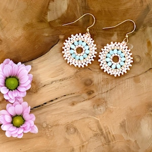 May include: A pair of gold hoop earrings with a beaded design. The beads are in shades of pink and light blue, creating a circular pattern. The earrings are on a wooden surface.
