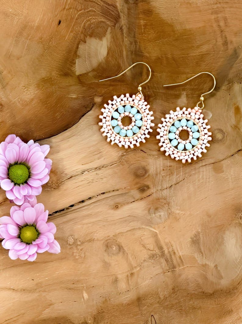 May include: A pair of gold hoop earrings with a beaded design. The beads are in shades of pink and light blue, creating a circular pattern. The earrings are on a wooden surface.