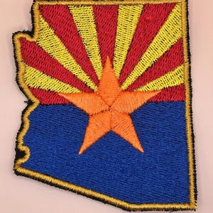 May include: Embroidered patch in the shape of Arizona, featuring the state flag design. The patch has a blue base with a large orange star in the center. Above the blue is a red and yellow sunburst design. The patch is outlined in gold.