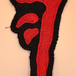 Può includere: Toppa ricamata con un simbolo rosso stilizzato su sfondo nero. Il simbolo ha un design astratto unico con un contorno spesso. La toppa è probabilmente per abbigliamento o accessori.