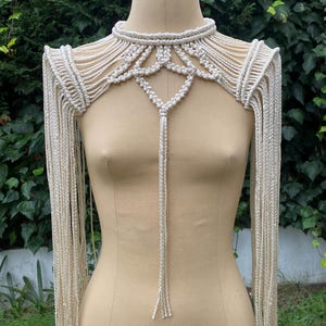 Op de afbeelding: Een wit macramé schouderstuk met een lange hanger en franjes. Het stuk is gemaakt van een dik koord en heeft een geometrisch ontwerp.