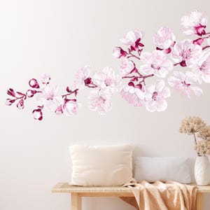 Op de afbeelding: Een witte muur met een roze en witte bloemen sticker. De sticker toont een tak met kersenbloesems met roze bloemen en bordeauxrode stelen. De sticker is geplaatst boven een houten bank met een wit kussen en een beige deken.