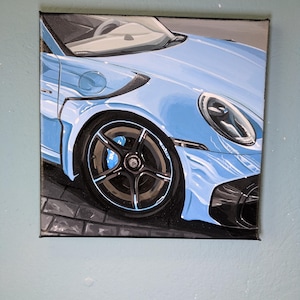 May include: A square canvas painting depicting the front quarter view of a light blue sports car. The artwork features a detailed black five-spoke wheel with blue brake calipers, a sleek headlight, and dark carbon fiber accents on the body.
