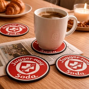May include: A white coffee mug rests on a red and white coaster with the text "Jugger-nog Soda". Other coasters with the same design are on a wooden table, with a newspaper and a plate of croissants.