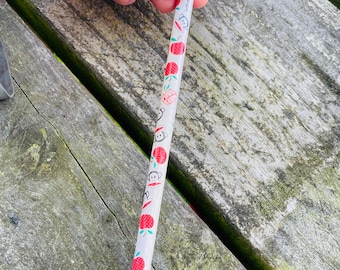 May include: A white pencil decorated with red apple and monkey illustrations. The pencil has a pink eraser and the word "Toffun" printed on it. The pencil is held against a weathered wooden surface.